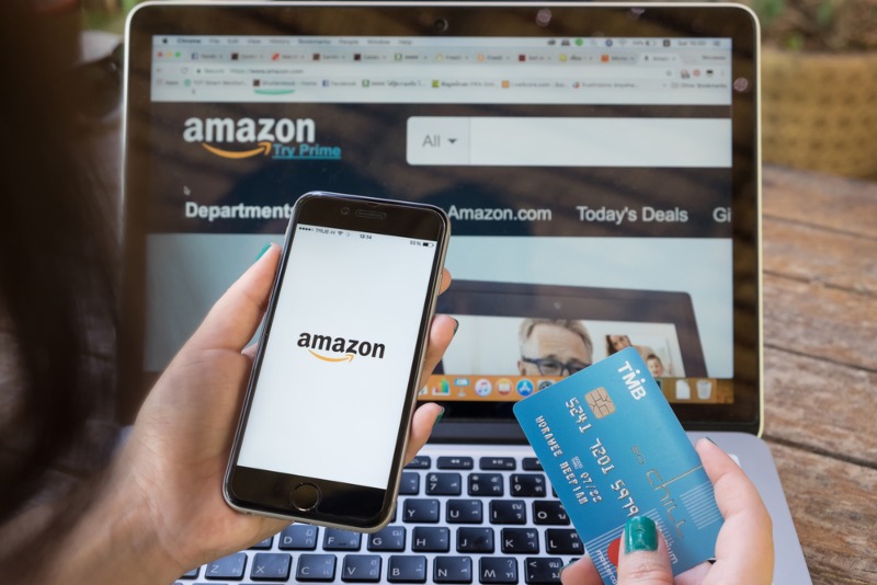 shot of a person looking at their phone and laptop showing the Amazon logo while holding a credit card